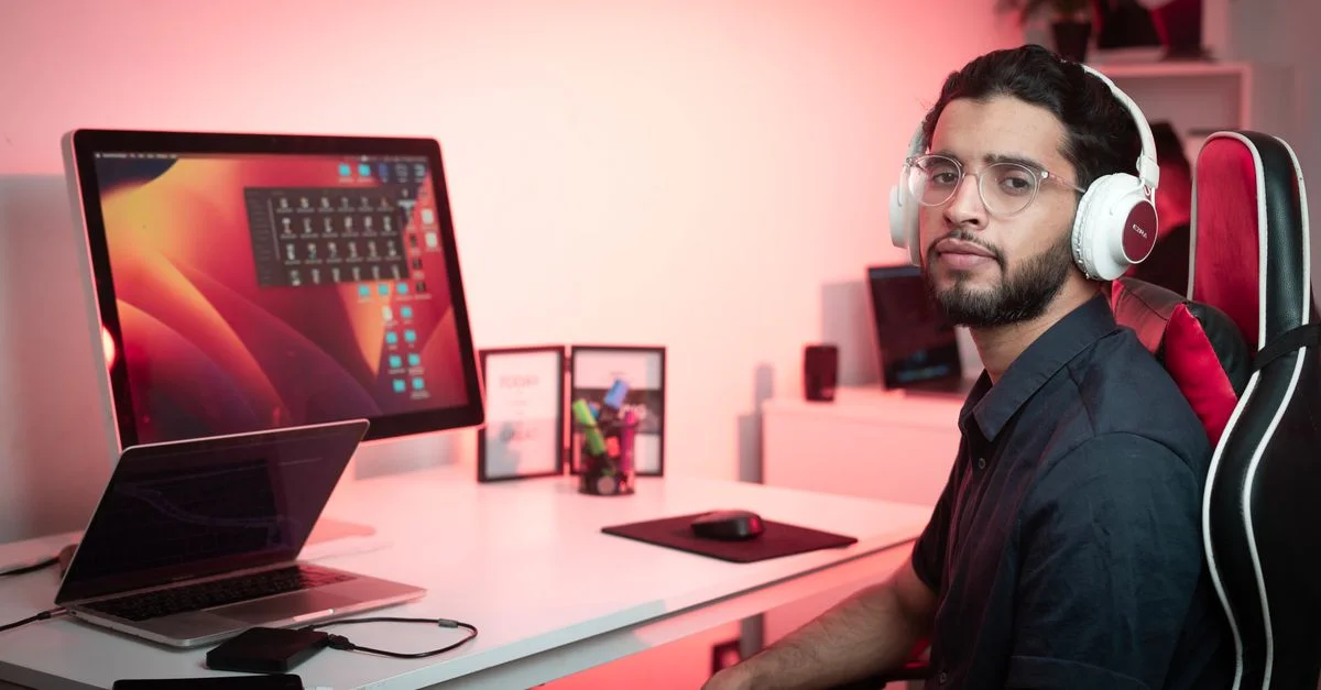 Jeune homme avec des écouteurs travaillant à un bureau avec des écrans doubles dans un cadre de bureau moderne.